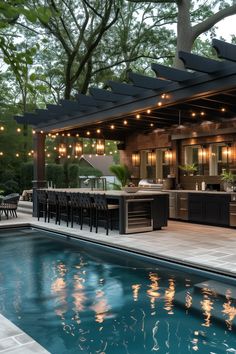 an outdoor kitchen and dining area next to a swimming pool with lights hanging from the ceiling