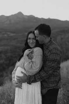 a man and woman hugging each other while standing in a field with mountains in the background