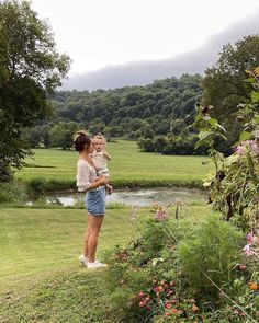 a woman holding a child in her arms while standing on the grass near flowers and trees