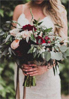 a woman holding a bouquet of flowers and greenery on her wedding day in the woods