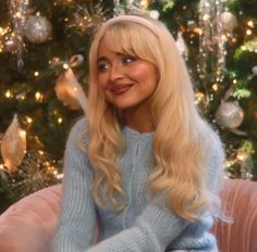 a blonde woman sitting in front of a christmas tree wearing a blue sweater and smiling