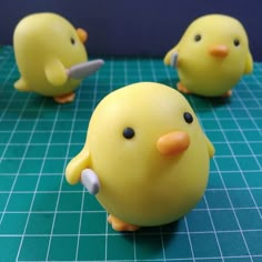 three yellow plastic birds sitting on top of a green table next to each other and one has a knife in it's mouth