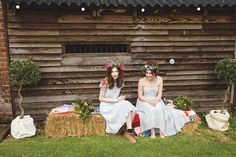 two young women sitting on hay bales in front of a wooden building with lights strung from the roof