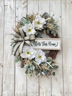 a wreath with white flowers and greenery hanging on a wooden wall next to a sign that says, bess this home