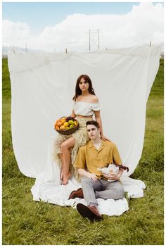 a man and woman sitting on top of a blanket with fruit in front of them