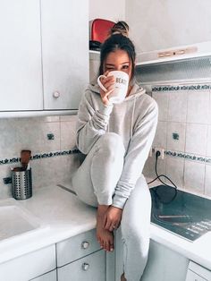 a woman sitting on top of a kitchen counter drinking from a mug while wearing sweatpants