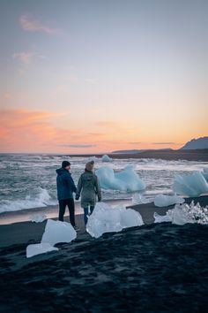 two people walking on the beach with icebergs in the water at sunset or dawn