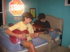 two boys sitting on a bed playing guitars