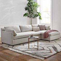 a living room with white walls and wood flooring, a large sectional couch in front of a potted plant