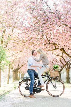 a man and woman riding on the back of a bicycle under cherry blossom trees in full bloom