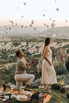 two people standing on top of a hill with hot air balloons in the sky