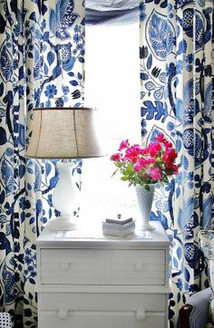 a blue and white room with flowers on the dresser in front of the window,