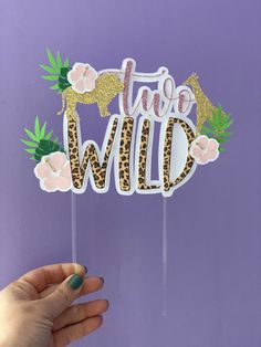 a hand holding up a cake topper with the word wild on it and flowers