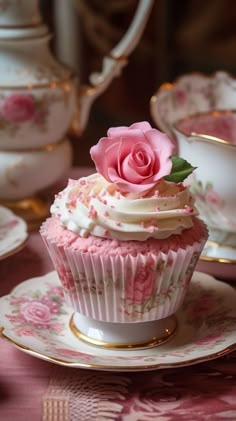 a cupcake on a saucer with a pink rose on top and tea cups in the background