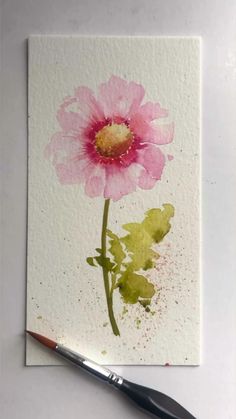 a pink flower is sitting on top of a piece of paper next to a paintbrush