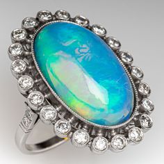 This gorgeous ring is centered with one (1) oval cabochon cut natural crystal opal and set into a bezel setting. The opal is bordered with twenty-two (22), bezel set, round brilliant cut diamonds, The top face of the shank is accented with three (3), bead set, round brilliant cut diamonds on each side. The gemstones are bordered with milgrain edging. The ring measures 29.1mm at the top, rises 10.6mm above the finger, tapering to 1.8mm wide and 1.1mm thick at the base of the shank. This ring is currently a size 6. Oval Cabochon Opal Ring Fine Jewelry, Fine Jewelry Oval Cabochon Opal Ring, Oval Cabochon Opal Ring In Fine Jewelry, Formal Oval Cabochon Opal Ring Fine Jewelry, Opal Diamond Ring Oval Cabochon, Diamond Opal Ring Oval Cabochon, Luxury Oval Opal Ring With 17 Jewels, Formal Opal Oval Cabochon Ring, Elegant Multi-stone Opal Ring With Oval Cabochon