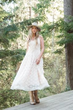 a woman in a white dress and hat standing on a wooden deck next to trees