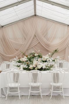 a table set up with white chairs and flowers on it in front of a curtained wall