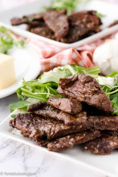 two plates with meat and greens on them sitting on a marble counter top next to cheese