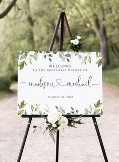 a welcome sign with greenery and white flowers on it is standing in the middle of a dirt road