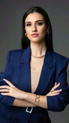 a woman with her arms crossed wearing a blue suit and gold bracelets, standing in front of a gray background