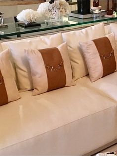 a white couch with some brown and white pillows on it's back, sitting in front of a glass coffee table