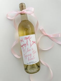 a bottle of wine with a pink ribbon around it on a white surface, and the label says she's tying the knot