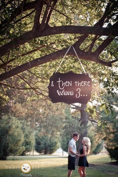 a couple standing under a tree with a sign hanging from it's trunk that says i turn three years old