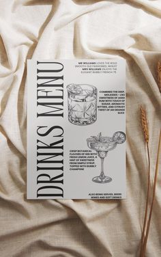 an open book with drinks on it sitting on top of a white bed sheet next to some dry grass