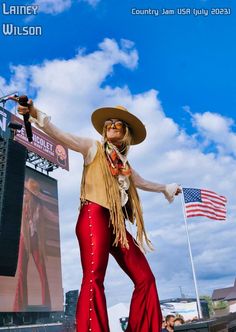 a woman in red pants and a cowboy hat is performing on stage with an american flag behind her