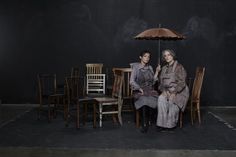 two women sitting under an umbrella on top of a black floor next to wooden chairs