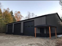 two garages in the middle of a dirt lot with trees around them and one has a black roof