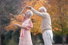 an older couple dancing together in the park on a fall day with trees turning orange