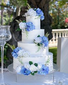 a white wedding cake with blue flowers and greenery on the top is surrounded by wine glasses