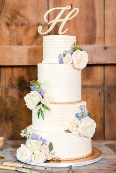 a white wedding cake with flowers and the letter h on top is sitting on a wooden table