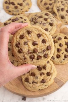 a person holding a chocolate chip cookie on top of a wooden board with other cookies