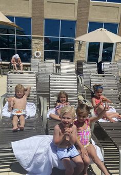 several children sitting on lounge chairs in front of a building with sunbathers and umbrellas