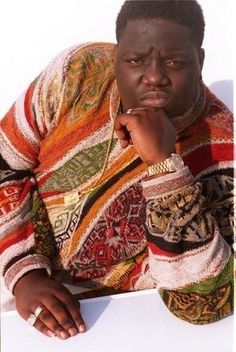 a man sitting down with his hand on his chin and looking at the camera while wearing a multicolored sweater