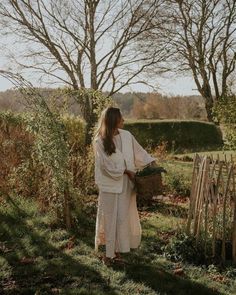 a woman standing in the grass next to a wooden fence and some trees with leaves on it