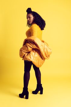 a woman in a yellow jacket and black tights is standing against a yellow background