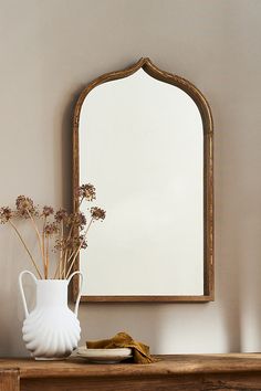 a white vase with flowers and a mirror on a wooden shelf in front of a wall