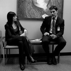 two people sitting on chairs in front of a large painting and one is talking to the other