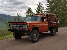 an orange truck with logs in the back driving down a dirt road next to trees