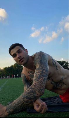 a man with tattoos on his arm and chest sitting on the ground in front of a soccer field