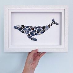 a hand holding up a white framed artwork with blue and black pebbles in it's shape