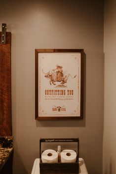 two rolls of toilet paper sitting on top of a wooden box in front of a framed poster