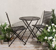 an outdoor table and chairs with flowers on the ground in front of a white wall