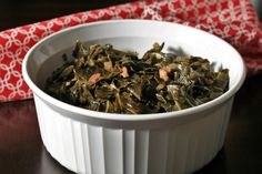 a white bowl filled with cooked greens on top of a wooden table next to a red and white napkin