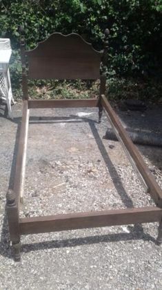 an old wooden bed frame sitting in the middle of a gravel ground next to some bushes