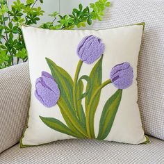 a white pillow with purple crocheted flowers on it sitting on a couch next to a potted plant